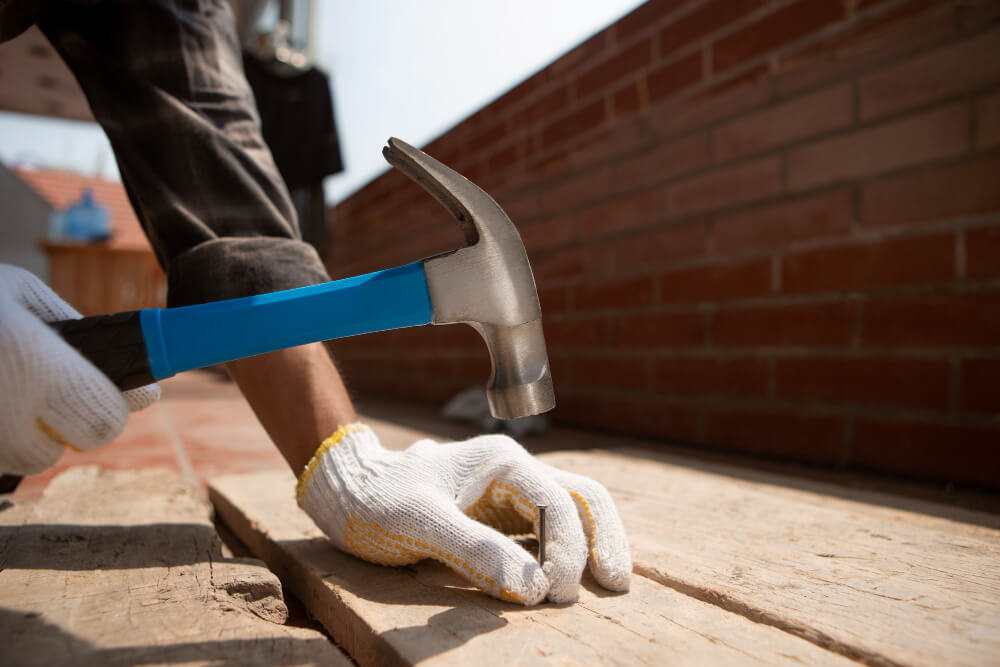 Expert roofer with gloved hands using hammer to drive a nail into wood
